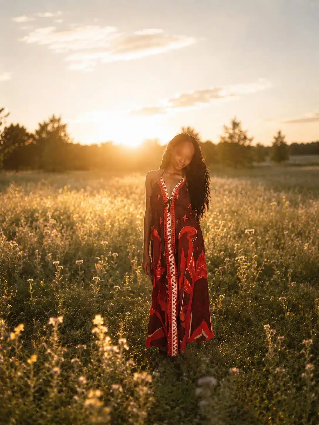 Eine Frau in einem langen, rot gemusterten Kleid steht in einem sonnenbeschienenen Feld mit Wildblumen bei Sonnenuntergang, mit Bäumen und einem leuchtenden Himmel im Hintergrund.