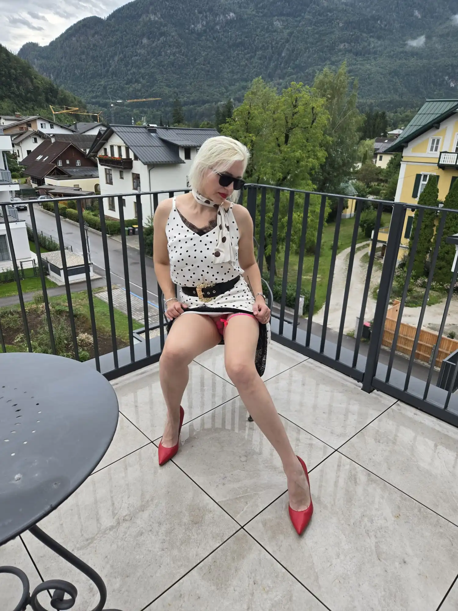 Eine Frau namens Eva, die ein weißes Kleid mit Punkten und rote Pumps trägt, sitzt auf einem Balkonstuhl und blickt nach unten. Im Hintergrund sind Häuser, Bäume und Berge zu sehen, im Vordergrund steht ein runder Tisch.